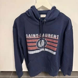 Marinblå Saint Laurent hoodie M - Snygg marinblå hoodie från Saint Laurent med tryck i vitt och rött framtill. Tröjan har huva med snörning, stor magficka och ribbade muddar. Tillverkad i mjuk bomull, perfekt för chill dagar. Klassisk streetstil med coolt grafiskt tryck.