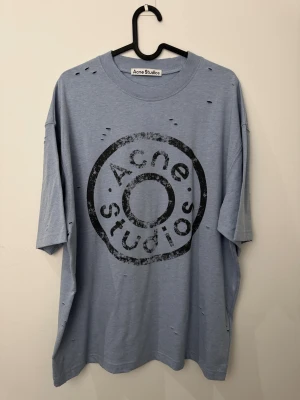 Blå Acne Studios t-shirt med jeans knapp tryck storlek M - Snygg ljusblå t-shirt från Acne Studios med stort, slitet svart logotryck på bröstet. Oversized passform och korta ärmar. Materialet är mjuk bomull blandat med polyester. Distressed detaljer med små hål ger en cool, edgy vibe.