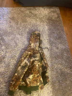 Carhartt camouflage vintage jacka med huva - Säljer en fet camouflage vindjacka från Carhartt med huva och gröna muddar. Jackan har dragkedja framtill, två fickor och är fodrad på insidan. Perfekt för dig som vill sticka ut med en streetig look och gillar outdoor-vibbar.