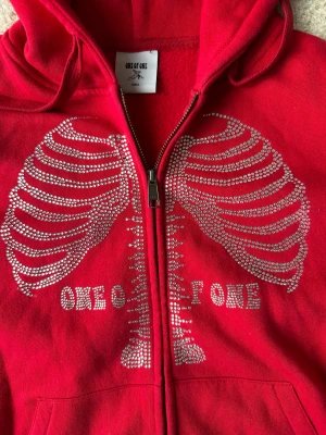 One Of One Hoodie   - Säljer en röd hoodie från One of One i storlek small. Tröjan har huva, dragkedja och fickor framtill. På bröstet finns ett coolt strassmönster som föreställer revben och texten 'One of One'. Perfekt för dig som gillar unika detaljer och statement-plagg.
