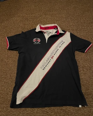 Paul & Shark marinblå pikéskjorta - Snygg marinblå pikéskjorta från Paul & Shark med vit och röd diagonal detalj över framsidan, broderad logga och tryck på bröstet. Kragen är vit med röd insida och knappar framtill. Perfekt för en sportig och stilren look.