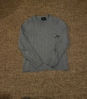 Grå kabelstickad tröja från bondelid - Grå kabelstickad tröja från bondelid med rund hals och långärmad passform. Tröjan har ett litet broderat emblem på bröstet och ribbade muddar vid ärmslut och nederkant. Perfekt för dig som gillar klassisk stil med en modern touch.