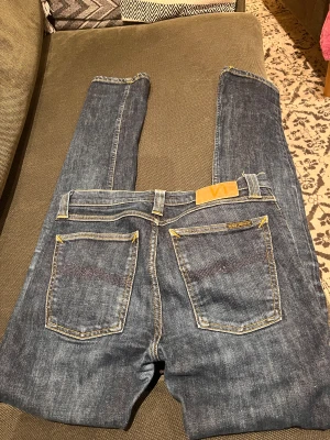 Mörkblå Nudie Jeans, strl 31/34 - Snygga mörkblå jeans från Nudie Jeans med klassisk femficksdesign och kontrasterande gula sömmar. Modellen har rak passform och är tillverkad i slitstarkt bomullsdenim. Perfekt för dig som gillar stilrena och tidlösa jeans.
