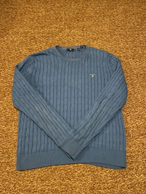 Blå kabelstickad tröja från Gant - Snygg blå kabelstickad tröja från Gant med rund hals och diskret logga på bröstet. Tröjan har långa ärmar och breda ribbade muddar vid ärmslut och nederkant. Perfekt för dig som gillar klassisk stil med en modern touch.