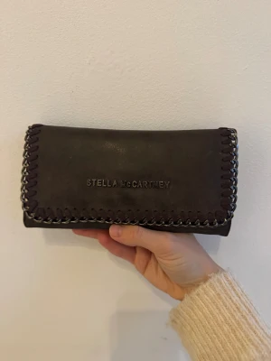 Stella McCartney Plånbok - Snygg brun plånbok från Stella McCartney med kedjedetaljer längs kanterna och logotyp framtill. Plånboken har en rektangulär form och är tillverkad i skinnliknande material med dekorativ söm runt om. Perfekt storlek för att ha med sig överallt. Nästan oanvänd! 
