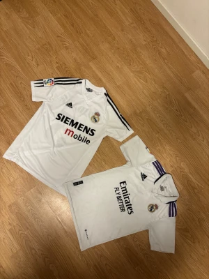 Real Madrid tröjor Zidane & Benzema - Två klassiska Real Madrid fotbollströjor från Adidas. Ena har tryck med Zidane #5 storlek M och Siemens Mobile sponsor, den andra har Benzema #9 storlek S och Emirates Fly Better sponsor. Båda är vita med svarta detaljer, korta ärmar och klubbmärke på bröstet. Perfekt för dig som älskar fotboll och samlar på legender.