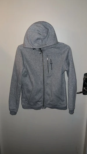 Grå hoodiejacka från Sail Racing - Säljer en grå hoodiejacka från Sail Racing med dragkedja och ficka på bröstet. Jackan har lång ärm, huva och svart logga på framsidan. Mjuk insida och sportig look, perfekt för chill dagar. Passar dig som gillar enkel och clean stil. Köpte den sommaren 2024 för 700 kr men har inte använt den sedan dess 