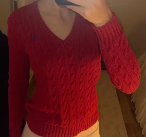 Röd stickad tröja från Ralph Lauren - Säljer en snygg röd kabelstickad tröja från Ralph Lauren med blå broderad logga på bröstet. Tröjan har v-ringad hals, långa ärmar och ribbade muddar. Perfekt för dig som gillar klassisk stil med en modern touch. Skriv om du har frågor! ÄKTA!