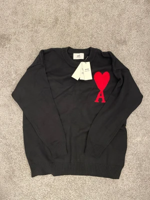 Svart stickad tröja från Ami - Svart stickad tröja från Ami Alexandre Mattiussi med röd hjärt- och A-logga på bröstet. Tröjan har rund hals, ribbade muddar och är långärmad. Materialet är mjuk ull och passformen är normal. Perfekt för dig som gillar stilrena detaljer och snygga loggor.