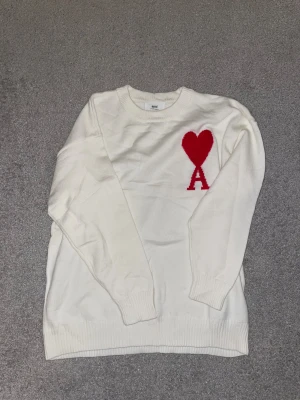 Vit stickad tröja från Ami Paris - Vit stickad tröja från Ami Paris med rött hjärta och bokstaven A broderat på bröstet. Rund hals, ribbade muddar och långärmad. Tröjan har en clean look med snygg kontrastdetalj framtill, perfekt för dig som gillar stilrena plagg med en twist.