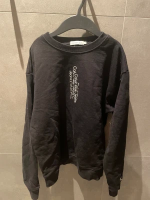 Svart sweatshirt från Calvin Klein Jeans - Svart sweatshirt från Calvin Klein Jeans med vit logotyp vertikalt på bröstet och liten CK-brodyr på ärmen. Tröjan har rund hals, ribbade muddar och är tillverkad i mjuk bomull. Perfekt för en chill och stilren look.