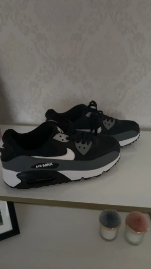 Nike Air Max 90 svart och grå - Säljer ett par stilrena Nike Air Max 90 sneakers i svart, grått och vitt. Skorna har klassisk snörning, rund tå och en synlig Air Max-sula för extra komfort. Materialet är en mix av syntet och skinn med coola detaljer och kontrasterande färger.