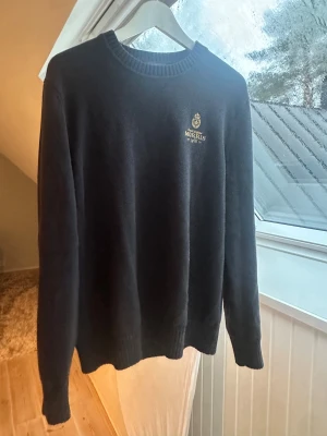 Mörkblå stickad tröja från Morris - Snygg mörkblå stickad tröja från Morris med broderad logga på bröstet. Tröjan har rund hals och ribbade muddar vid ärmslut och nederkant. 