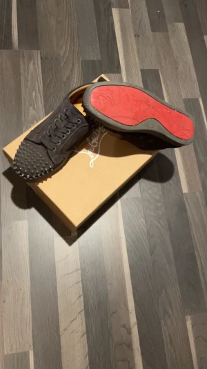 Svarta Louboutin sneakers med nitar Loubs byte - Säljer ett par svarta sneakers från Louboutin med ikoniska röda sulor och coola nitar över tån. Skorna har snörning och är tillverkade i skinn för en lyxig känsla. Perfekta för dig som vill sticka ut med edgy detaljer och exklusiv vibe. Byter