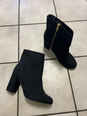 Svarta mocka boots med klack - Säljer ett par svarta boots i mjuk mocka-look med chunky klack och rund tå. Skorna har guldfärgade dragkedjor på sidan som snygg detalj. Perfekta till höst- och vinteroutfits för dig som vill ha en clean men ändå edgy stil.
