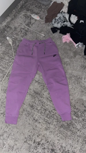 Lila mjukisbyxor från Nike - Säljer ett par lila mjukisbyxor från Nike med svart logga på låret. Byxorna har snörning i midjan med svartvita snören och muddar vid bensluten. Perfekta för chill dagar eller träning.