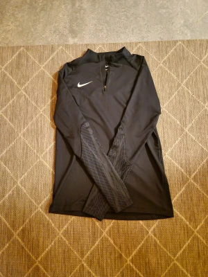 Svart Nike Dri-Fit half zip tröja - Svart långärmad tröja från Nike med Dri-Fit-teknologi och slim fit-passning. Snyggt mönstrade detaljer på ärmarna och en praktisk half zip framtill. Perfekt för träning och sportiga dagar, med klassisk Nike-logga på bröstet. Tyvärr finns det ett litet hål längst ner men syns knappt. Passar som S/M i storleken