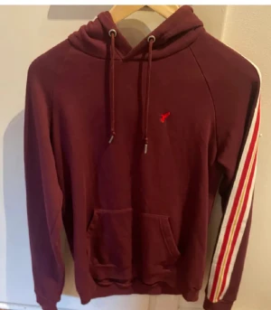 Vinröd hoodie med ränder på ärmarna - Säljer en vinröd hoodie från Lyle & Scott med klassisk huva och snörning. Tröjan har vita, orange och röda ränder längs ärmarna samt en liten röd logga på bröstet. Stor ficka framtill och ribbade muddar. Perfekt för chill dagar.
