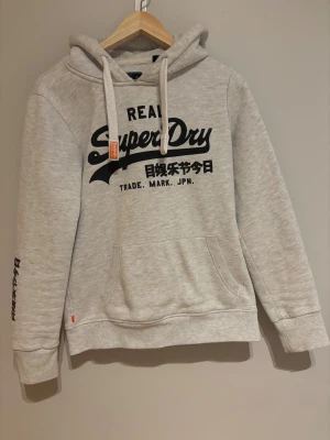Superdry hoodie grå - En stilren Superdry hoodie i färgen grå med svart tryck. Den är knappt använd och är i superbra skick! Det står storlek M i den men sitter mer som storlek S. Pris kan diskuteras! 