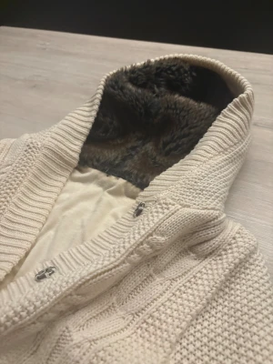 Beige stickad hoodie med päls - Mysig beige stickad hoodie med grov kabelstickning och ribbade muddar. Insidan av huvan är fodrad med mjuk päls i mörkbrunt, vilket gör den extra varm. Tröjan har två fickor framtill och snygga metallknappar vid halsen. Perfekt för kalla dagar. Storlek S/Xs