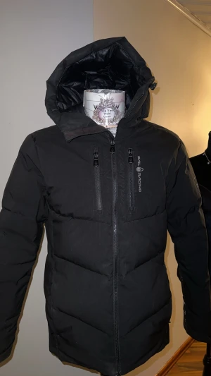 Svart dunjacka Sail Racing Antarctica - Svart dunjacka från Sail Racing Antarctica Expedition med huva och två bröstfickor med dragkedja. Jackan har snygga logodetaljer på bröstet och ärmen, samt en stilren, puffad design som håller dig varm. Perfekt för kalla vinterdagar och har en modern look. Jackan är S men passar M