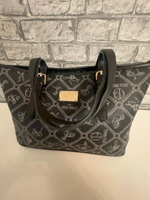 Svart handväska från Giulia Pieralli - Snygg svart handväska med silverfärgat rutmönster och detaljerade illustrationer. Väskan har två rejäla handtag, guldfärgad logoplatta framtill och är tillverkad i skinnimitation. Perfekt för dig som gillar stilrena accessoarer med unik design.