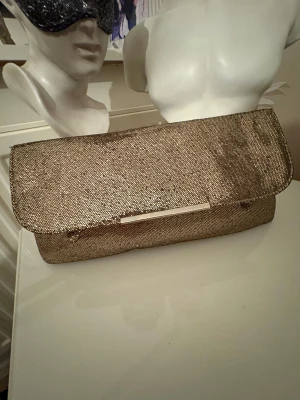 Glittrig guld kuvertväska med kedja - Snygg kuvertväska i glittrigt guld med metallkedja som axelrem. Väskan har ett stilrent metallspänne och magnetlås. Insidan är ljus med ett fack och dragkedja. Perfekt för att lyfta din outfit med lite extra bling.