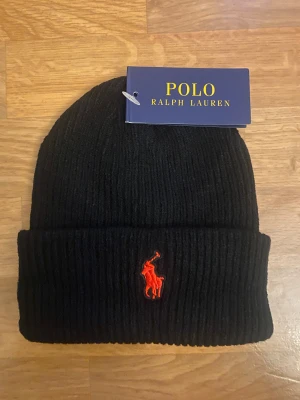 Polo Ralph Lauren Mössa - Svart ribbstickad mössa från Polo Ralph Lauren med klassisk röd broderad logga framtill. Snygg uppvikt kant och mjukt material som håller dig varm. Perfekt accessoar för kyliga dagar och enkel att matcha med din stil.