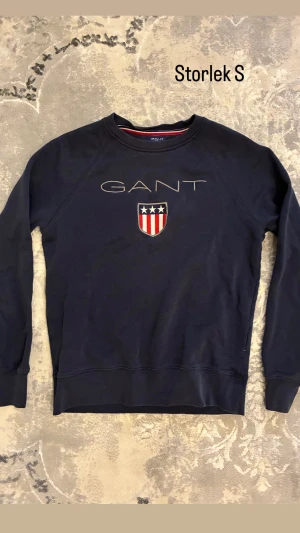 Mörkblå sweatshirt från GANT - Snygg mörkblå sweatshirt från GANT med broderad logga och emblem på bröstet. Tröjan har rund hals, långa ärmar och ribbade muddar vid ärmslut och nederkant. Perfekt för dig som gillar klassisk och sportig stil.