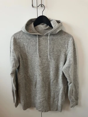 Grå stickad hoodie med huva - Mysig grå hoodie i stickat material med huva och snörning. Tröjan har långa ärmar och ribbade muddar vid ärmslut och nederkant. Perfekt för dig som gillar chill och enkel stil. Passar dig som vill ha en basic men ändå snygg look.