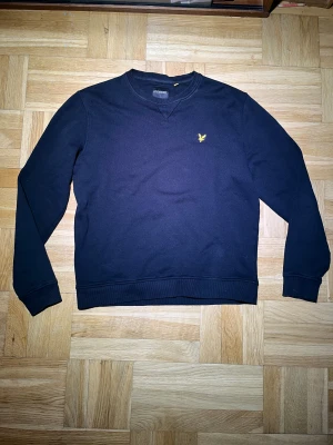 Svart sweatshirt från Lyle & Scott - Svart sweatshirt från Lyle & Scott med klassisk rund hals och gul broderad fågel på bröstet. Tröjan har ribbade muddar vid ärmslut och nederkant, och är tillverkad i mjuk bomull. Perfekt för chill dagar och enkel att matcha med jeans eller joggers.