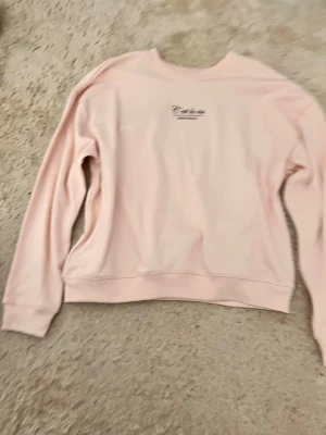 Ljusrosa sweatshirt  - Säljer en ljusrosa sweatshirt med diskret tryck på bröstet. Tröjan har rund hals, långa ärmar och ribbade muddar vid ärmslut och nederkant. Perfekt för chill dagar och enkel att matcha med jeans eller leggings.