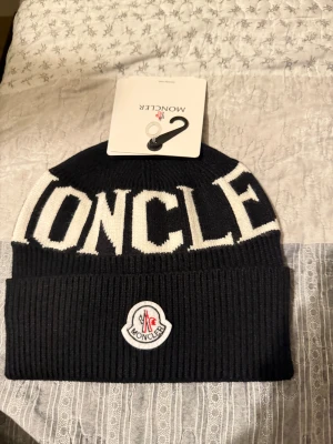 Svart och vit Moncler mössa - Snygg svart och vit mössa från Moncler med stor logotyp framtill och broderad patch på uppvikt kant. Stickad i mjukt material med ribbad struktur och tydlig Moncler-text i vitt. Perfekt för dig som vill ha en cool och exklusiv look under kalla dagar.