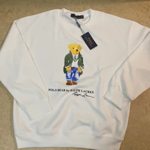 Vit sweatshirt Polo Bear Ralph Lauren - Vit sweatshirt från Polo Ralph Lauren med ikoniskt Polo Bear-tryck på framsidan. Tröjan har rund hals, långa ärmar och ribbade muddar. Materialet är mjuk bomull och passformen är normal. Perfekt för dig som gillar klassisk streetstyle med en twist.