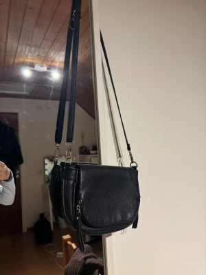 Svart axelväska i skinn med dragkedja - Svart axelväska i skinn med rundad form och silverfärgade detaljer. Väskan har både magnetlås och dragkedja, samt flera fack. Justerbar axelrem. 