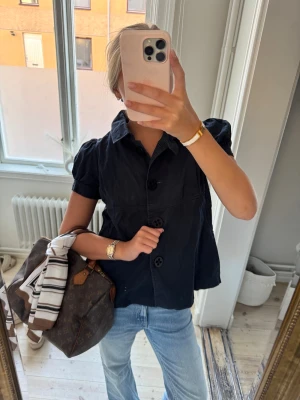 Svart blus med puffärm och stora knappar - Säljer en svart blus med korta puffärmar och stor krage. Blusen har tre stora knappar framtill och en lätt A-linjeformad siluett. Materialet känns som bomull och ger en avslappnad men ändå chic vibe. Perfekt för dig som gillar klassiska detaljer med en twist.