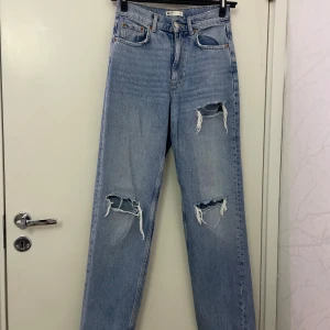 Blå ripped jeans från Gina Tricot - Snygga ljusblå jeans från Gina Tricot med slitna detaljer och hål på båda benen. Modellen är högmidjad och har raka ben med klassisk femficksdesign. Jeansen är tillverkade i bomull och har en cool vintagekänsla.