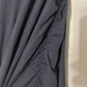 Mörkblå långklänning med rynkade detaljer - Säljer en stilren mörkblå långklänning med snygga rynkade detaljer längs ena sidan. Klänningen har en rak och enkel siluett som ger en clean look. Perfekt för dig som gillar minimalistisk stil och vill ha något som sticker ut med subtila detaljer.