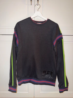 Svart MCQ sweatshirt med ränder - Cool svart sweatshirt från MCQ med gröna, blå och lila ränder längs ärmarna och nederkanten. Tröjan har lång ärm och ett MCQ-tryck på framsidan. Perfekt för dig som gillar streetwear och vill sticka ut med färgade detaljer.