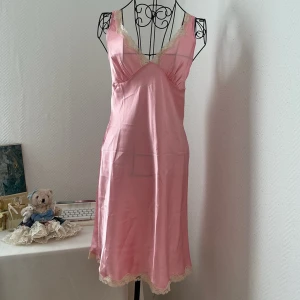 Rosa satinklänning från Victoria’s Secret - Supersöt rosa satinklänning med v-ringad hals och tunna axelband. Klänningen har krämvit spets längs urringning och nederkant som ger en romantisk vibe. Den täcker troligtvis inte knä om du är längre än 155cm. Storlek XS, passar 32-34. Dockan har storlek 34.