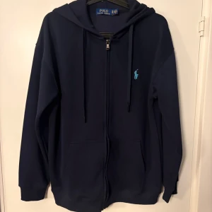  Ralph Lauren Zip-Up - Snygg marinblå hoodie från Polo Ralph Lauren med dragkedja och huva. Tröjan har långa ärmar, två fickor fram och den klassiska broderade loggan i ljusblått på bröstet. Perfekt för chill dagar och enkel att matcha med jeans eller joggers. Slim fit passar er som är runt M/L