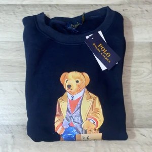 Mörkblå Polo Bear tröja Ralph Lauren - Snygg mörkblå sweatshirt från Polo Ralph Lauren med ikoniskt Polo Bear-tryck på bröstet. Tröjan har rund hals, långa ärmar och ribbade muddar. Materialet är mjuk bomull som känns skönt mot huden. Perfekt för dig som gillar klassisk streetstyle med en twist.
