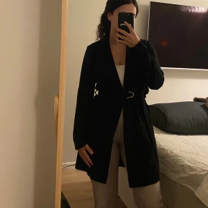 Svart kappa med bälte - Svart kappa med klassisk krage och bälte i midjan. Jackan har långa ärmar och en stilren, rak passform. Materialet känns mjukt och lätt, vilket gör den enkel att bära över olika outfits.