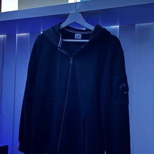Svart hoodie från C.P. Company - Svart hoodie från C.P. Company med dragkedja framtill, luva och den klassiska linsdetaljen på ärmen. Tröjan har två stora fickor fram och ribbade muddar. Storlek L, men passar mer M. Passar inte mig längre, så säljer nu denna.