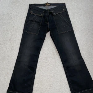 Svarta jeansbyxor med stora fickor - Svarta jeansbyxor från Voi Jeans med breda ben och stora, snedställda fickor framtill. Kontrastsydda sömmar och detaljerade bakfickor med knapp. Klassisk låg midja och bootcut-snitt för en cool och avslappnad look.