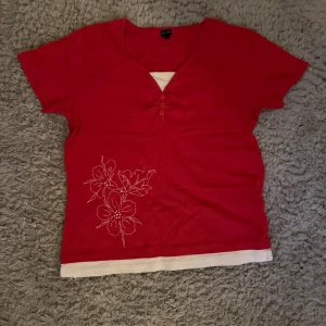 Röd v-ringad topp med blommor - Säljer en röd v-ringad topp från Zara med korta ärmar och dekorativa knappar vid halsen. Toppen har ett vitt lager nedtill och ett snyggt blommigt broderi på ena sidan. Perfekt för dig som gillar färg och detaljer!