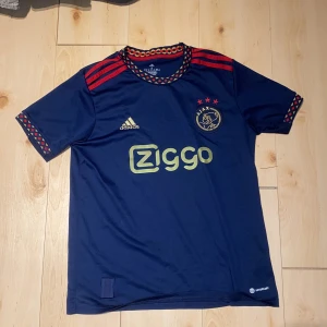 Ajax blå fotbollströja Adidas S - Ajax fotbollströja i mörkblått från Adidas med röda och gula detaljer. Tröjan har korta ärmar, röda ränder på axlarna och mönstrad krage och ärmslut. Ajax-logga och Ziggo-sponsor på bröstet. Tillverkad i lätt och ventilerande polyester.