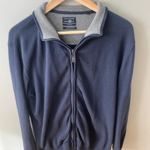 Marinblå kofta från A.W. Dunmore - Kofta i marinblått från A.W. Dunmore med hel dragkedja och kontrasterande grå ribbad krage. Tillverkad i 100% bomull, långärmad och med klassisk passform. Perfekt för lager-på-lager och snygg till jeans eller chinos.