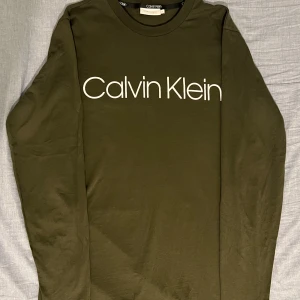 Olivgrön långärmad tröja Calvin Klein - Snygg olivgrön långärmad tröja från Calvin Klein med stor vit logga framtill. Tröjan har rund halsringning och är tillverkad i mjuk bomull, perfekt för en clean och stilren look.