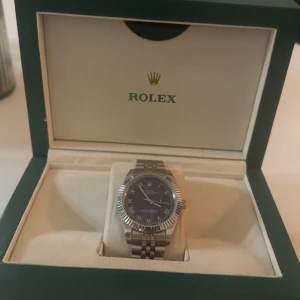 Rollex Datejustt 41mm azuro roman dial  - Exklusiv Rollex Datejustt med blå urtavla, romerska siffror och klassisk räfflad bezel. Klockan har en elegant silverfärgad länk i rostfritt stål och levereras i originalbox med internationellt garantikort. En tidlös accessoar från ett av världens mest ikoniska klockmärken.